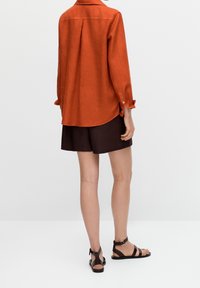 Chemise en lin orange rouille avec des poignets à bouton, associée à un short marron foncé. La chemise présente un col décontracté et des fentes sur les côtés. Sandales noires incluses.