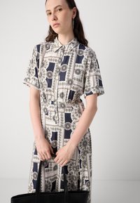 Short-sleeve dress featuring a geometric print in navy, cream, and green, with a buttoned front and a relaxed fit. Black bag held in hands.