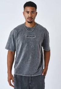 Camiseta de algodón gris con efecto ácido y mangas cortas, con un logotipo rectangular en el centro y un ajuste relajado. Cuello ajustable con detalles en las costuras.