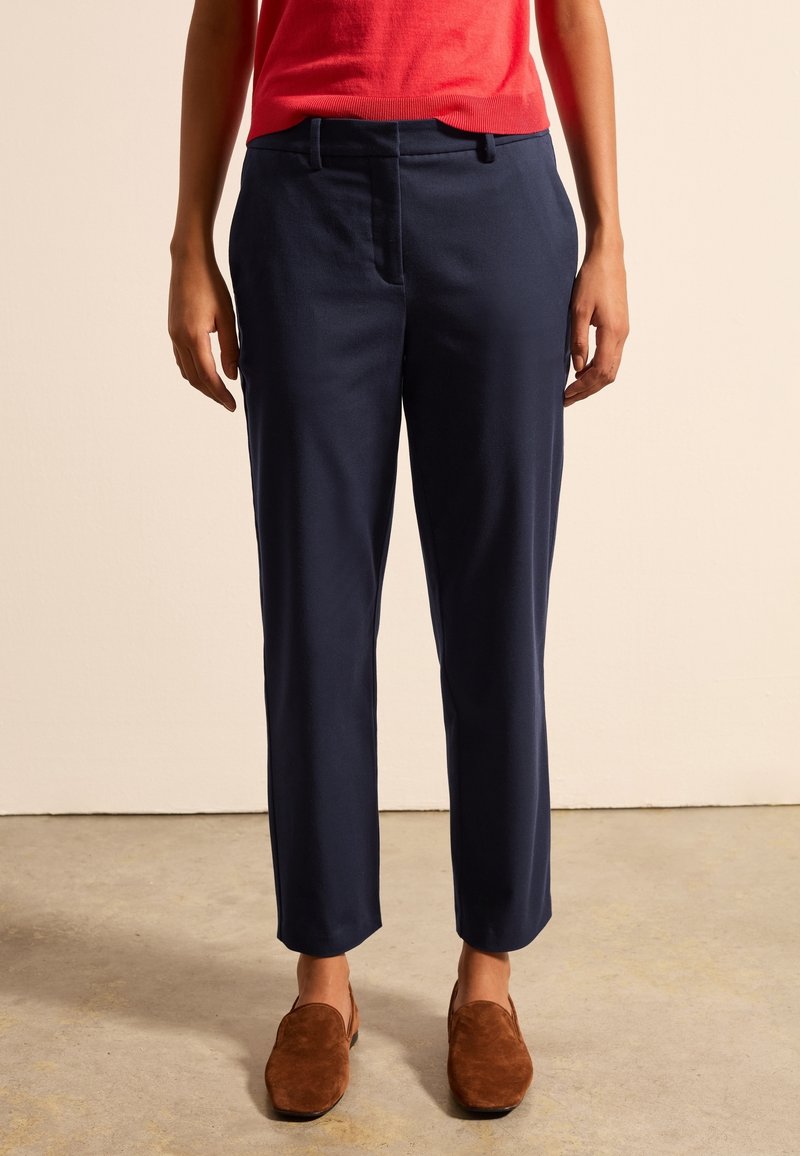 Granatowe, dopasowane spodnie o prostych nogawkach, z zakładkami, kieszeniami bocznymi i gładką teksturą; noszone z brązowymi zamszowymi butami.