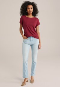 Punane lühikeste varrukatega tops, millel on zikk-zakk muster, koos helesiniste sirgete pükste ja beežide plokknähtud kingadega. Ühtlane taust.