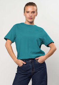 Femme portant un t-shirt turquoise à manches courtes et un jean bleu foncé, debout les mains dans les poches devant un fond clair uni.