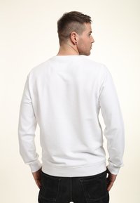 Man met kort haar en oorbellen, gekleed in een eenvoudige witte sweatshirt en zwarte spijkerbroeken, met zijn rug naar de camera gekeerd tegen een lichte achtergrond.