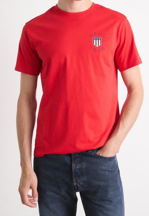 Man wearing a red short-sleeve t-shirt with a small GANT 1949 logo on the left chest and dark blue jeans.