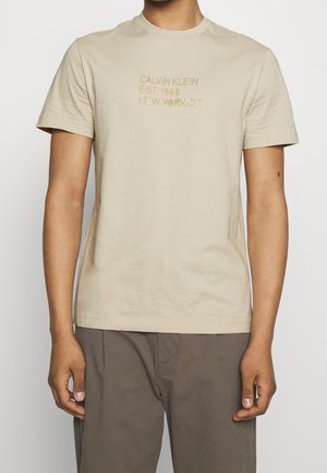 Beiges Baumwoll-T-Shirt mit rundem Ausschnitt und kurzen Ärmeln, mit erhabenem goldfarbenem Schriftzug "CALVIN KLEIN EST. 1968 NEW YORK CITY."