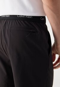 Juodi šortai su plokščiu užpakaliniu kišeniu ir elastinga liemene, ant kurios matomas "Calvin Klein" logotipas. Audinys atrodo lengvas ir glotnus.
