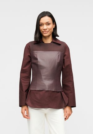 Μπορντό μακρυμάνικη μπλούζα με δομημένο δερμάτινο κορσέ, υφασμένο επάνω μέρος και βολάν στο τελείωμα, φορεμένη με λευκό παντελόνι.