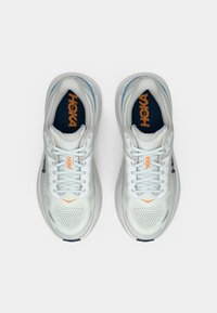 Tênis de atletismo em malha branca com detalhes em azul, bico arredondado, colarinho acolchoado e sola texturizada. Logo da HOKA em laranja na língua e nas laterais.