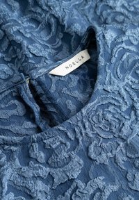 Niebieska teksturowana tkanina z wypukłymi kwiatowymi wzorami i gładkim wykończeniem; posiada metkę z napisem "NOELLA". Wzór obejmuje okrągły dekolt.