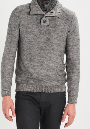 Mann trägt einen grauen Strickpullover mit geknöpftem Stehkragen und schwarze Hose, steht mit entspannten Armen an den Seiten.