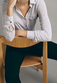Hellgrauhe Hemdbluse mit langen Ärmeln, ausgestattet mit zwei Brusttaschen und einer subtilen Textur, kombiniert mit schwarzen, eng anliegenden Hosen. Ein Holzstuhl ist sichtbar.