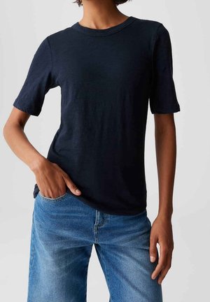 Personne portant un t-shirt noir uni à manches courtes et un jean bleu, avec une main dans la poche et un fond neutre.