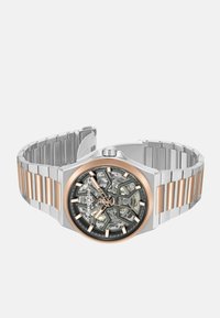 Srebrny i różowozłoty zegarek z ażurową tarczą, metalową bransoletą z naprzemiennie szczotkowanymi i polerowanymi ogniwami oraz okrągłą kopertą.