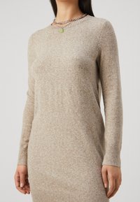 Robe-pull beige en maille avec manches longues, encolure arrondie, poignets côtelés et une texture subtile sur toute la surface. Accent notable avec un collier chaîne.