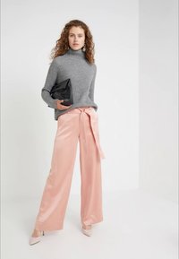 Vrouw staat in een grijze geribbelde coltrui, roze satijnen brede broek met een tailleband, huidskleurige hakken en houdt een zwarte clutch in haar hand.
