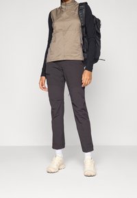 Lekki beżowy kamizelka na zamek z kontrastującymi ciemnymi rękawami, połączona z ciemnymi spodniami i beżowymi butami sportowymi. Czarny plecak dopełnia stylizację.