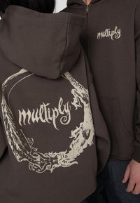Brun zip-up hoodie med et stort kontrasterende grafisk cirkulært design og ordet "multiply" i en dekorativ skrift på ryggen.