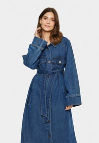 Džínsová šaty s dlhými rukávmi, predným zapínaním na gombíky a viazaním v páse. Má dve náprsné vrecká a modrú pranie s kontrastným šitím.