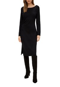 Schwarzes langärmeliges Kleid mit Rundhalsausschnitt, seitlicher Raffung und Schlitz. Getragen mit hohen schwarzen Wildlederstiefeln, die eine figurbetonte Silhouette betonen.