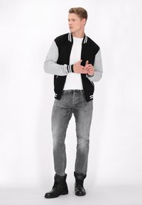 Jachetă bomber neagră și gri cu manșete în dungi, purtată peste un tricou alb. Blugi gri și cizme negre cu șireturi completează ținuta.