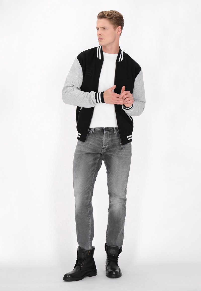 Schwarze und graue Bomberjacke mit gestreiften Bündchen, getragen über einem weißen T-Shirt. Graue Jeans und schwarze Schnürstiefel runden den Look ab.