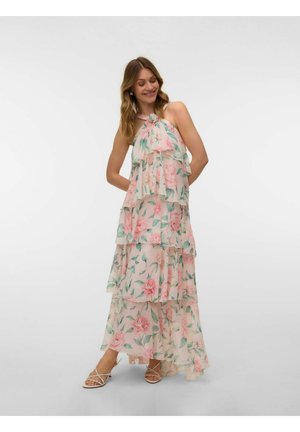 Žena nosí bezrukávové vrstvené kvetinové maxi šaty s ružovými kvetmi a zelenými listami, v kombinácii s béžovými sandálmi na podpätku so remienkami, usmieva sa s rukami za chrbtom.