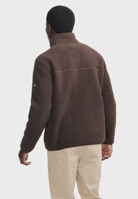 Brun fleecepullover med høj krave, tekstureret overflade og sidelommer, båret over lyse beige bukser. Minimal branding på ærmerne.