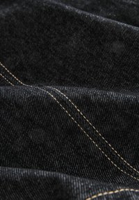 Tissu en denim noir avec une surface texturée, présentant des coutures contrastantes de couleur marron clair et des motifs de tissage diagonal visibles.