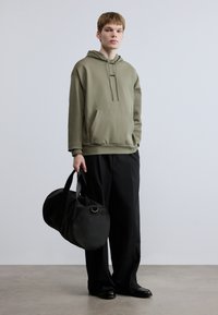 Λαδί φούτερ με κουκούλα και τσέπη τύπου καγκουρό, μαύρο λογότυπο στο στήθος, σε συνδυασμό με φαρδιά μαύρη παντελόνια και μεταφέροντας μια στρογγυλή μαύρη τσάντα duffel.