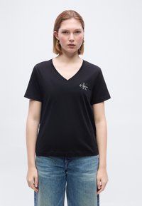 Γυναίκα με κοντά ξανθά μαλλιά που φοράει χρυσούς κρίκους σκουλαρίκια, μαύρο T-shirt Calvin Klein με V λαιμόκοψη και μπλε τζιν, στέκεται μπροστά σε απλό φόντο.