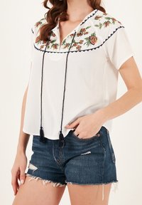 Chemisier blanc à manches courtes avec broderie florale au niveau du col, comportant des accents bleu marine et des pompons, associé à un short en jean.