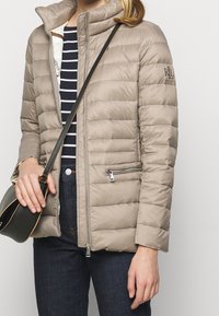 Taupefärgad vadderad jacka med hög krage, quiltad design, fickor med dragkedja och glansig textur, bars över en randig skjorta och mörka jeans.