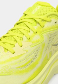 Helle gelbe Sportschuhe mit einem Obermaterial aus Mesh, dicker Sohle und flachen Schnürsenkeln. Verfügt über Branding und strukturierte Muster für Grip und Designdetails.