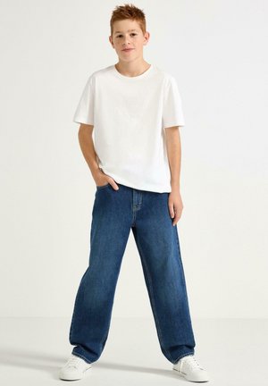 Vit bomullströja med korta ärmar, kombinerad med vida blå denimjeans. Modellen har vita sneakers och står framför en neutral bakgrund.
