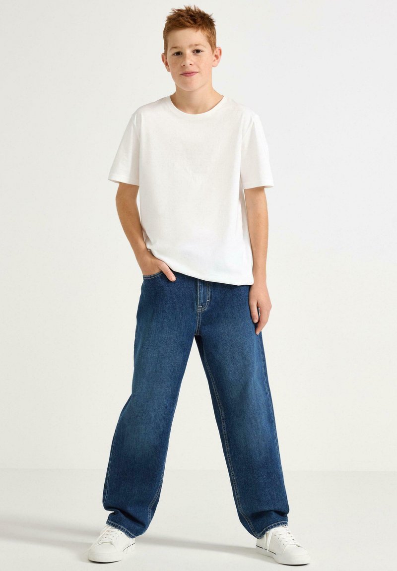 Vit bomullströja med korta ärmar, kombinerad med vida blå denimjeans. Modellen har vita sneakers och står framför en neutral bakgrund.
