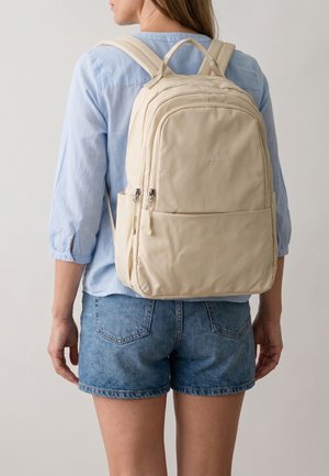 Kvinde iført lyseblå skjorte og denimshorts med en beige Venezia-rygsæk med flere lynlåslommer på ryggen.
