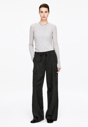 Woman standing wearing a light gray fitted long-sleeve top, black wide-leg cargo pants with drawstring, black shoes, and a silver necklace.