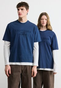 Δύο άτομα που φορούν μπλε σκούρα πουλόβερ "Tommy Jeans" πάνω από λευκά μακρυμάνικα και καφέ παντελόνια, στέκονται μπροστά σε ένα απλό φωτεινό φόντο.