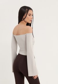Samantha apakšējā daļā gaismas pelēka auduma off-shoulder blūze ar garām zvana piedurknēm; frontē ir teksturēts ručings, apvienots ar pieguļošām brūnām biksēm.