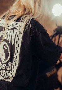 Czarny t-shirt z dużym nadrukiem graficznym w białym kolorze, z kratkową ramką i abstrakcyjnymi wzorami. Materiał wydaje się miękki i ma luźny krój.