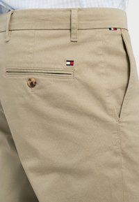 Beige trousers in a textured fabric with two back pockets. Features small logo accents in red, white, and blue near the waistband.