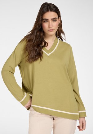Donna con capelli castani lunghi che indossa un maglione a V verde oliva con righe bianche e pantaloni beige, in posa con una mano sul fianco.