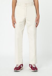 Mann in gerade geschnittenen, cremefarbenenHosen und burgunderfarbenen New Balance Sneakers mit weißen Schnürsenkeln vor einem weißen Hintergrund.