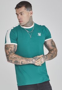 Maglietta verde con dettagli bianchi sulle spalle e sui bordi delle maniche. Presenta un piccolo logo bianco sul petto. Texture liscia, design aderente.
