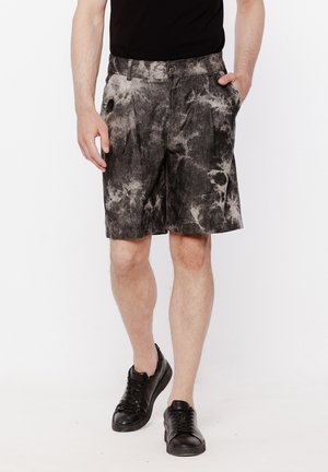 Mężczyzna noszący czarne i szare szorty tie-dye, czarną koszulkę i czarne trampki, stojący z rękami w kieszeniach na białym tle.