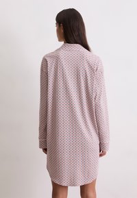 Femme aux longs cheveux noirs portant une robe à manches longues, de longueur genou, avec un petit motif géométrique, vue de dos sur un fond uni.