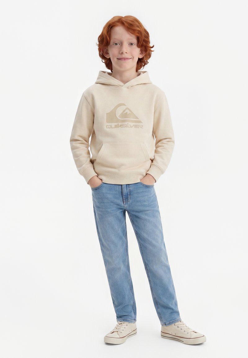 Κοκκινομάλλο αγόρι με μπεζ φούτερ Quiksilver, ανοιχτόχρωμα τζιν και άσπρα αθλητικά παπούτσια στέκεται με τα χέρια στις τσέπες σε λευκό φόντο.