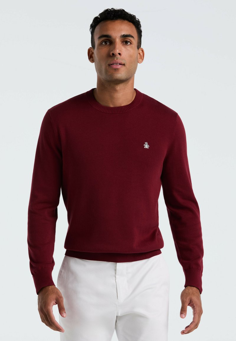 Bordeaux brei sweater met een ronde hals, lange mouwen en een subtiel logo op de borst; gedragen met witte broek. Gladde textuur.