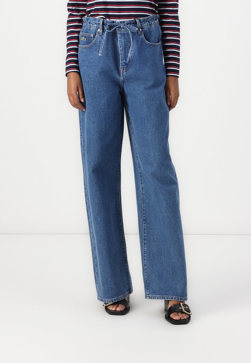 Lett blå, vide denimjeans med høy midje, frontlommer og et knytebelte-detalj. Har merkbare nagler og sømmer.