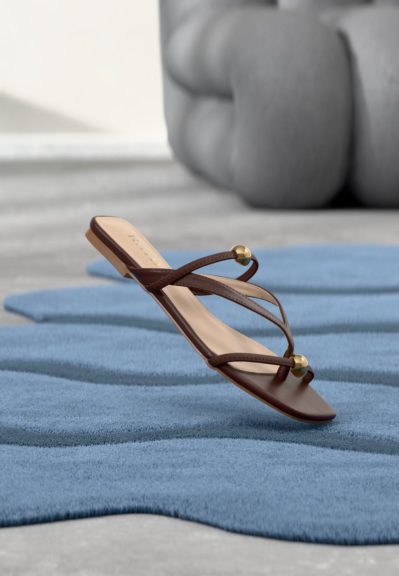 Καφέ επίπεδη σανδάλι με λεπτές λουρίδες και χρυσά χάντρες που ακουμπούν σε ένα υφασμένο μπλε χαλί, με ένα μεγάλο γκρι αφηρημένο γλυπτό στο φόντο.